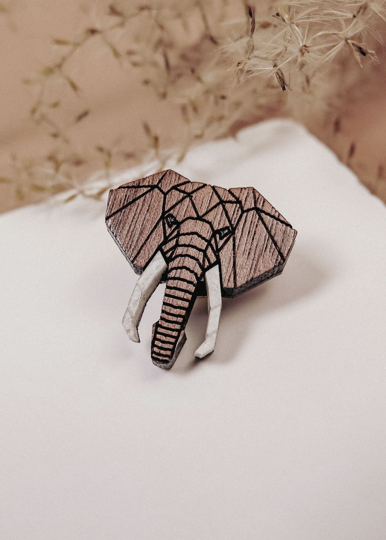 A handcrafted Elephant Pin shaped like an elephant’s head, featuring a striking geometric design, rests on a white surface with delicate dried flowers in the background.