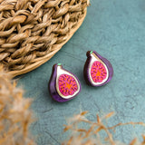 A pair of fig-shaped, hand-painted earrings with purple and pink detailing are displayed on a green surface next to a woven basket. Blurred beige plant stems are visible in the foreground.