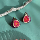 A pair of deep purple earrings, hand-painted to resemble figs with vibrant red centers, rest on a textured teal surface beside a white branch and a pink glass dish.