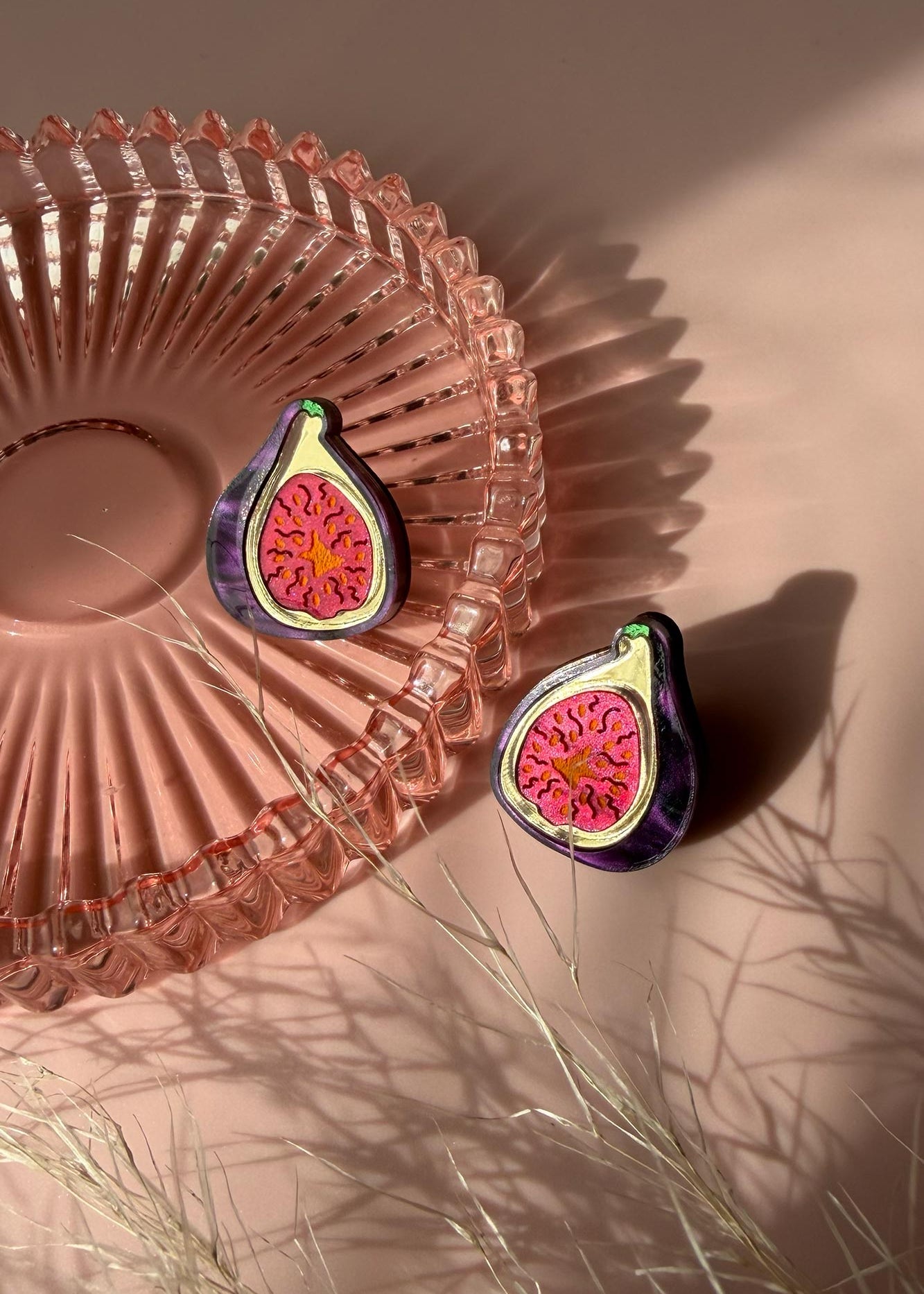 Two fig-shaped laser-cut studs with vibrant pink and deep purple detailing rest on a pink glass dish, casting shadows on a pale pink surface. Some delicate, dried grass lies nearby, adding texture to the scene.