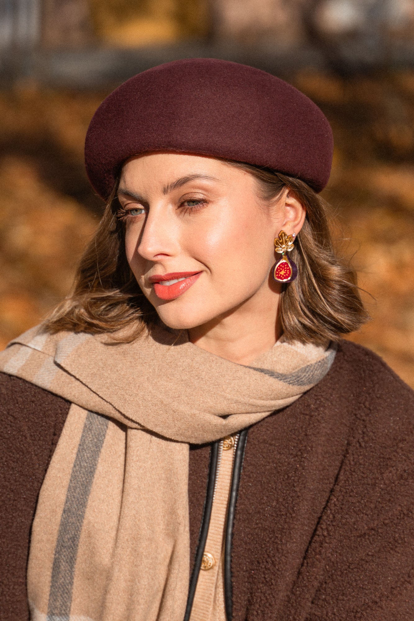 A woman wearing a maroon beret, beige and brown scarf, and matching coat smiles softly outdoors. She has light makeup and wears handcrafted Fig Earrings with gold mirror leaves. The background is blurred with autumn colors.