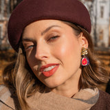 A woman wearing a burgundy beret and a beige scarf poses outdoors. She wears red lipstick, handcrafted fig earrings, and a matching pin. Autumn trees and a building create a scenic background.