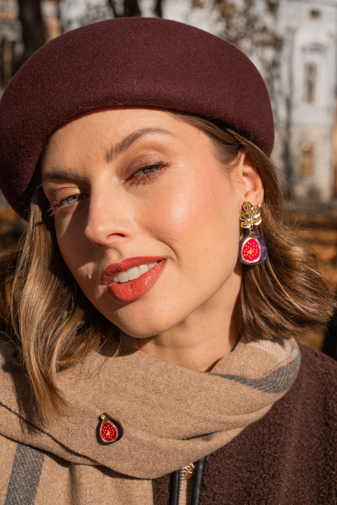 A woman wearing a burgundy beret and a beige scarf poses outdoors. She wears red lipstick, handcrafted fig earrings, and a matching pin. Autumn trees and a building create a scenic background.