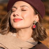A woman with light brown hair wearing a burgundy beret, beige scarf, and handcrafted fig earrings smiles with her eyes closed in warm sunlight, with a blurred autumn background.