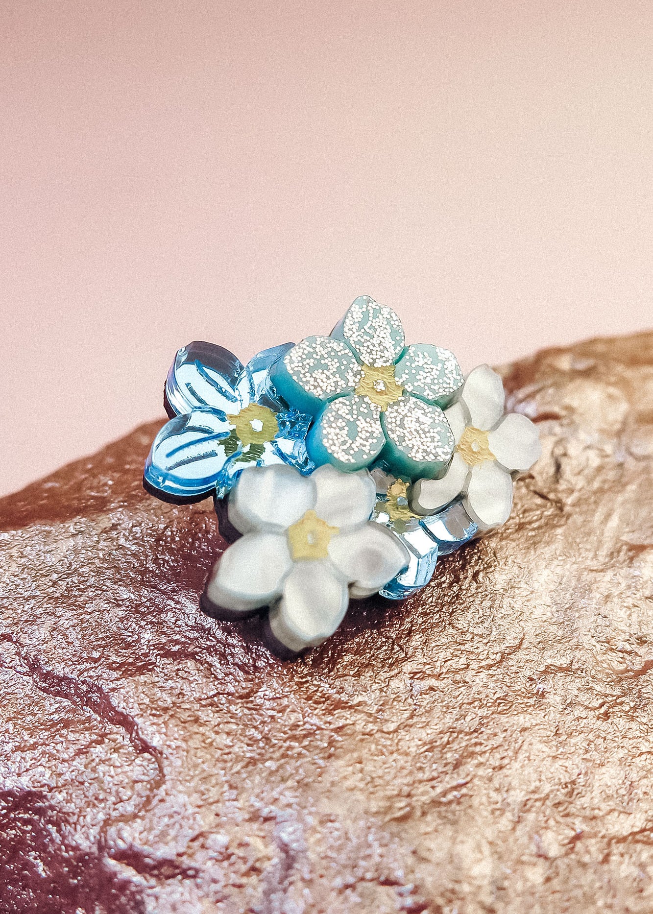 A cluster of delicate, handmade Forget Me Not Flower lapel pins with pastel blue and white petals and yellow centers rests on a textured rock against a soft pink background.