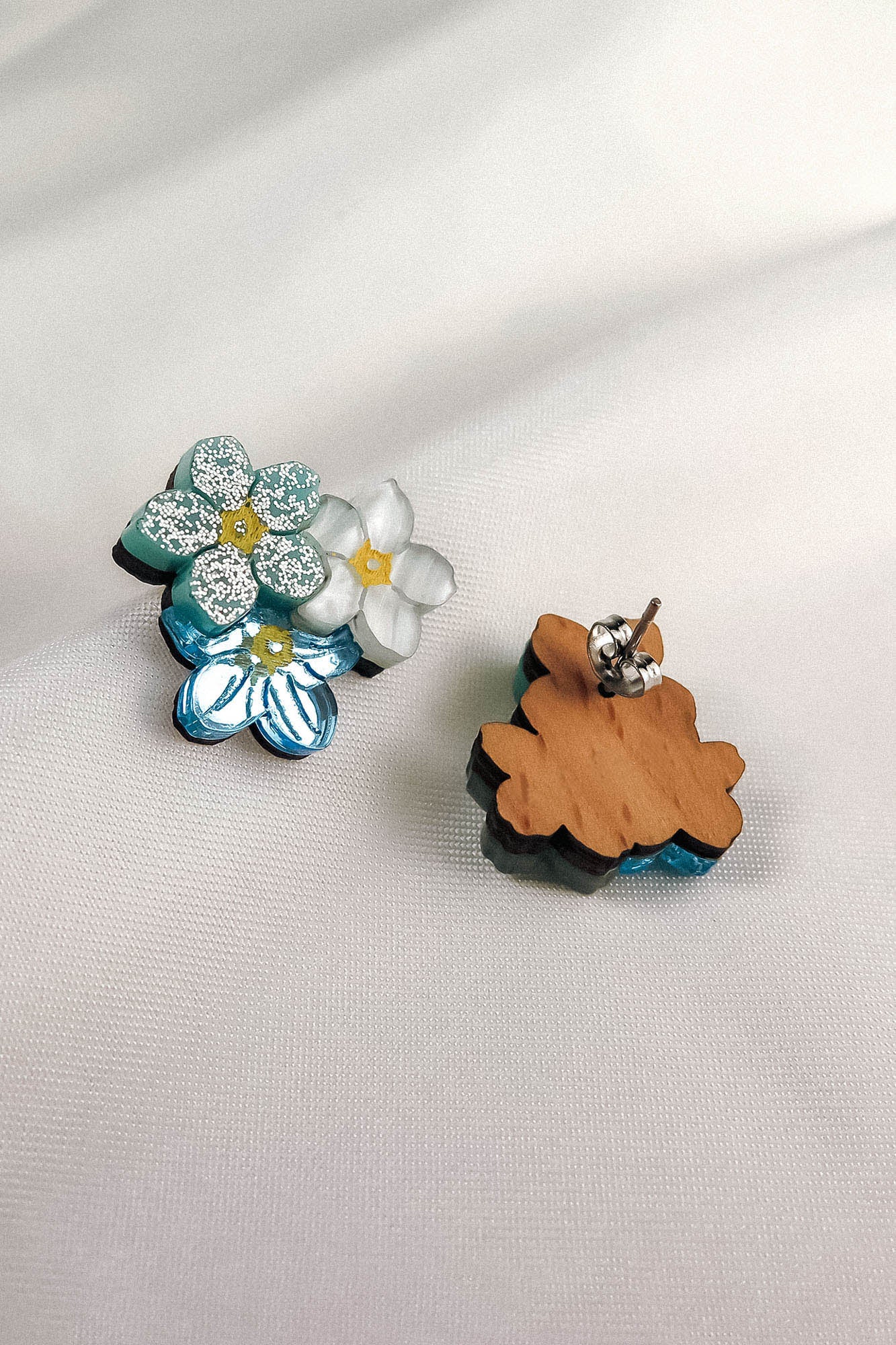 Two dainty floral earrings shaped like flowers rest on white fabric. One earring shows blue and white glitter details, while the other reveals the wooden back and metal post—these are the charming Forget Me Not Studs.