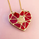 A handcrafted gold chain necklace featuring the Fragment of Love Necklace’s polygon heart pendant, formed from red stained glass outlined in gold, with a central gold plate etched with an arrow, set against a soft pink background.