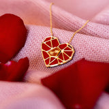 The handcrafted Fragment of Love Necklace features a heart pendant with a red, mosaic-like design, resting on soft pink fabric and surrounded by scattered red rose petals.