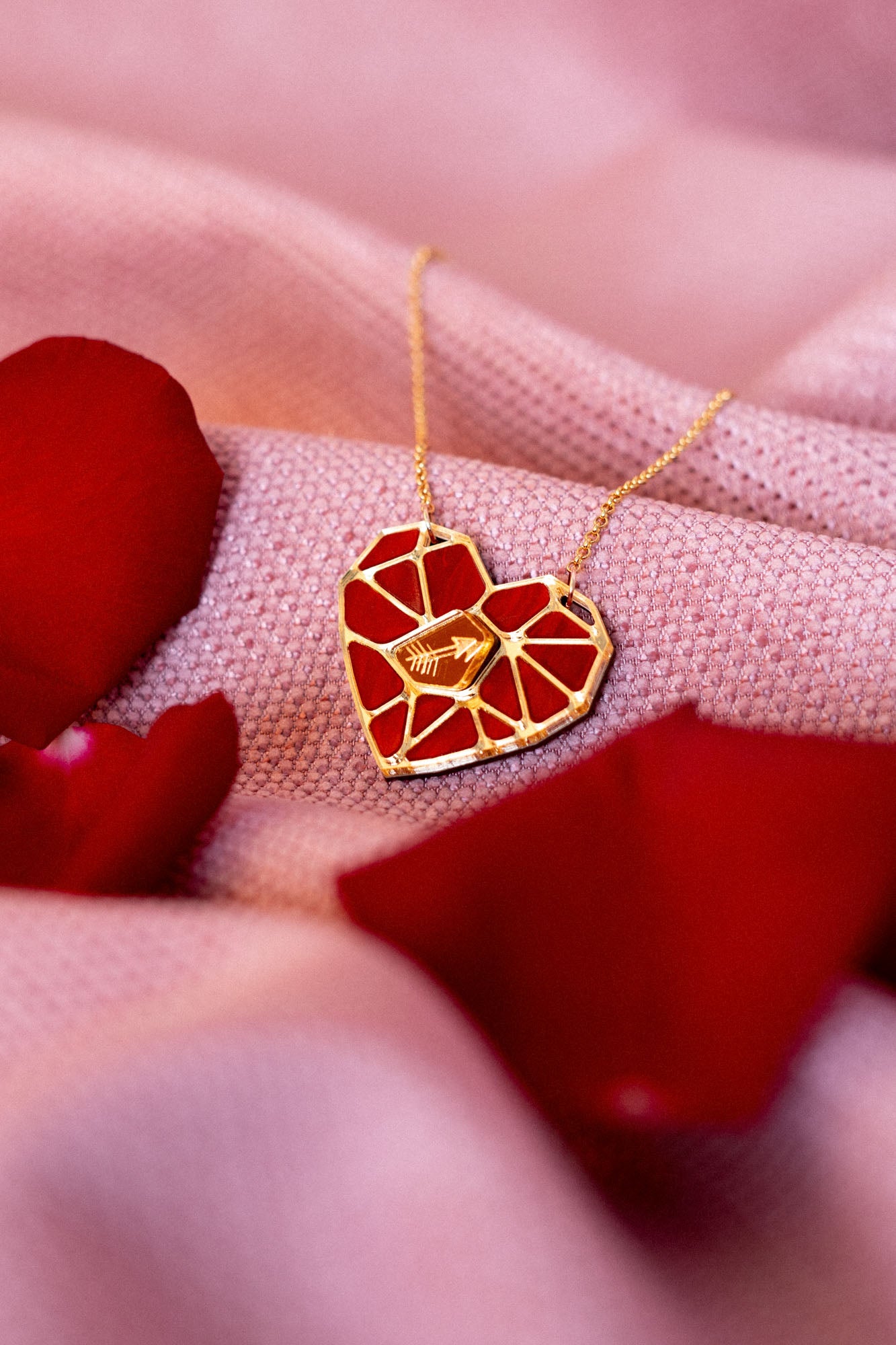 The handcrafted Fragment of Love Necklace features a heart pendant with a red, mosaic-like design, resting on soft pink fabric and surrounded by scattered red rose petals.