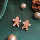 Two gingerbread man earrings rest on green fabric, surrounded by sparkling gold ornaments and a pinecone, making these festive accessories the perfect Christmas jewelry for any holiday look.