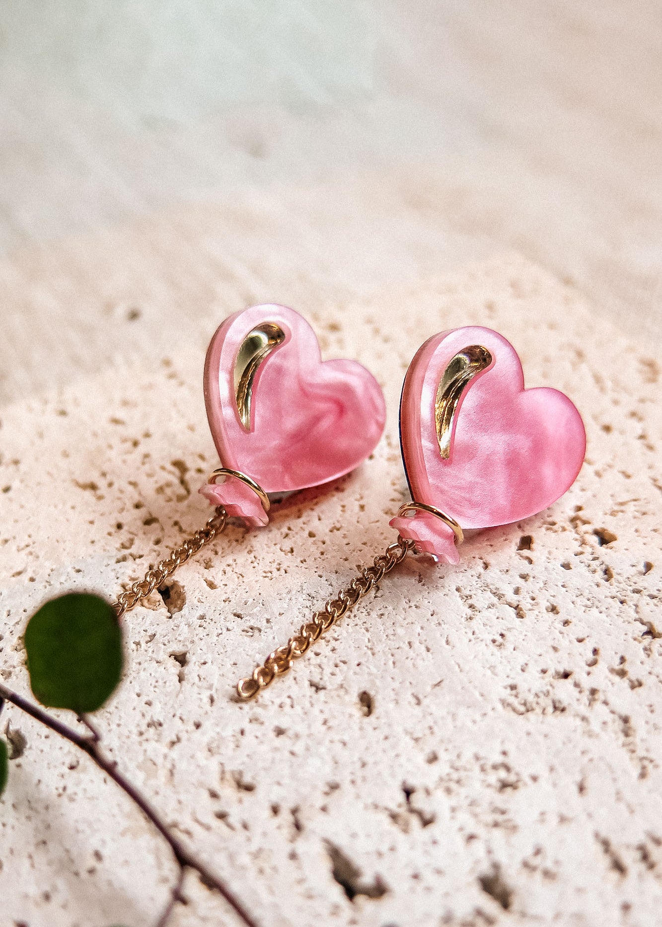 A pair of pink Heart Balloon Studs with gold accents and dangling gold chains, displayed on a textured, light-colored stone surface. A small green plant is visible in the lower left corner, completing this vibrant style.