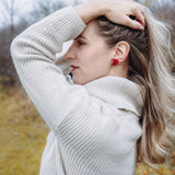 A woman in a cream-colored sweater stands outdoors, facing sideways with one hand holding back her long blonde hair. She wears playful Heart Balloon Studs, adding a romantic jewelry touch to her look. The background is blurry with trees and grass.