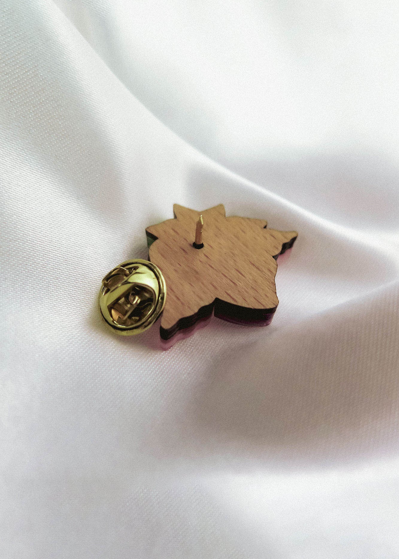 A handmade wooden lapel pin shaped like a maple leaf with a gold-colored clasp is resting on smooth white fabric.