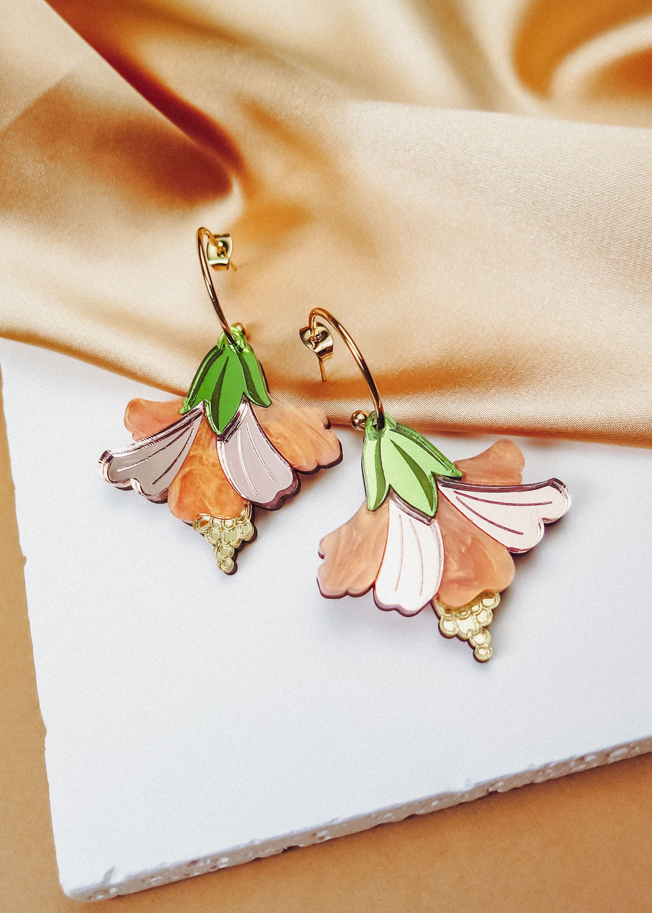 A pair of handcrafted Hibiscus Earrings with gold hoops, featuring pink, peach, and green petal designs, displayed on a white textured surface with beige satin fabric in the background.