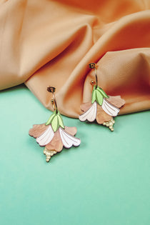 A pair of handcrafted Hibiscus Earrings, shaped like pastel-colored flowers with gold accents, are displayed on a pastel blue surface with peach-colored fabric in the background.