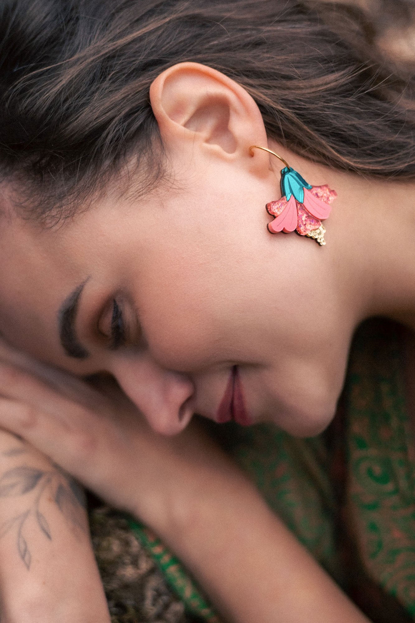 A woman with long brown hair lies on her side outdoors, displaying vibrant Hibiscus Earrings and a botanical tattoo on her arm. She is wearing a green patterned garment and appears calm and relaxed, making a tropical statement.