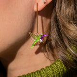 Close-up of a person’s ear wearing long, colorful handcrafted Hummingbird Earrings and a small stud earring. The person has light brown hair and is wearing a green knit sweater, showcasing sustainable craftsmanship.