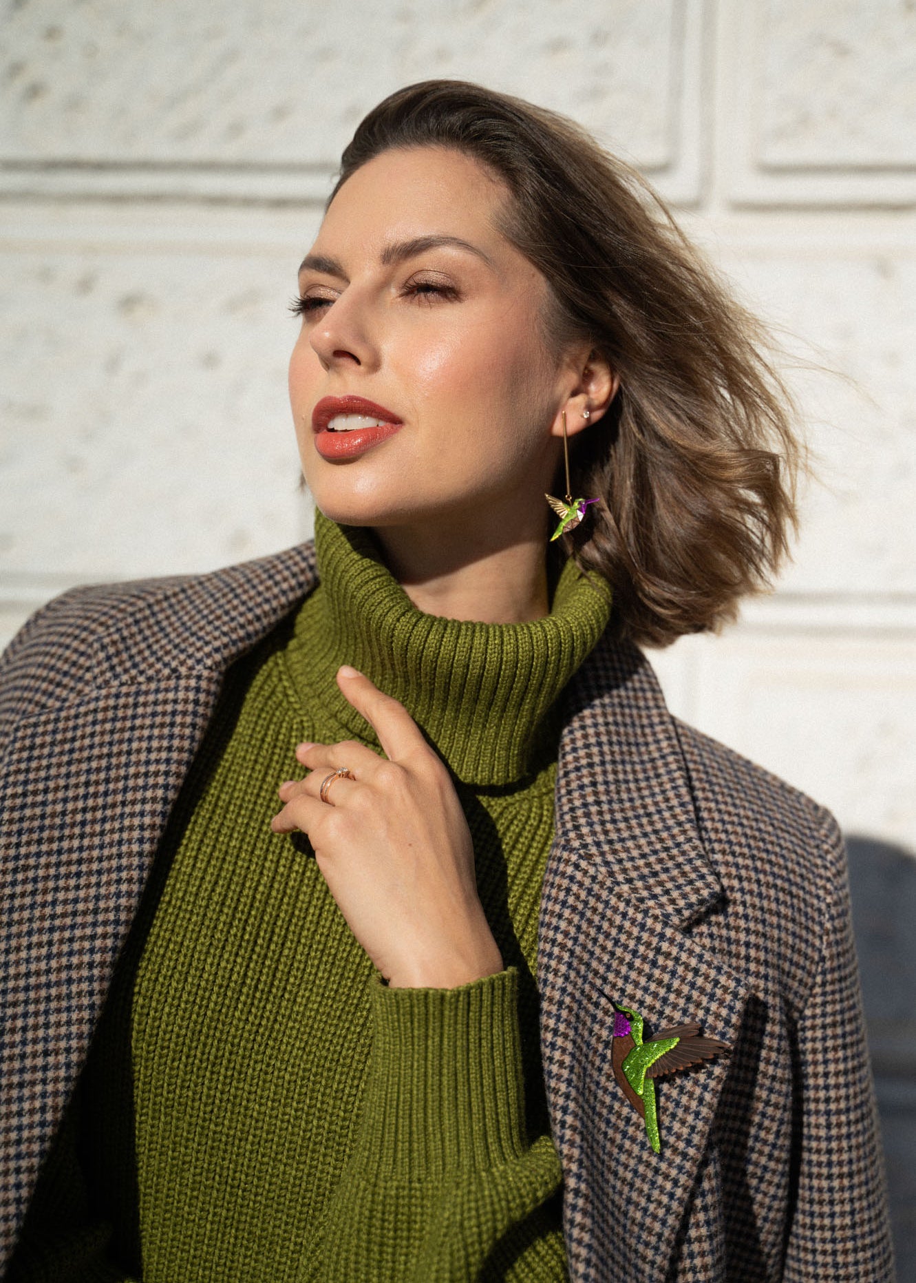 Woman wearing a green turtleneck sweater and plaid coat, accessorized with handcrafted Hummingbird Earrings and a brooch, poses outdoors against a light textured wall, touching her collar and looking off to the side.