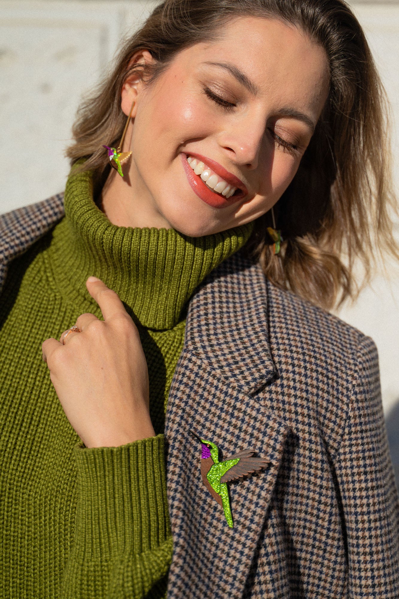 A smiling woman in a green turtleneck and plaid jacket stands against a light wall, wearing handcrafted Hummingbird Earrings and matching lapel jewelry. Sunlight highlights her face as she closes her eyes, radiating joy and sustainable craftsmanship.