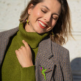 A smiling woman in a green turtleneck and plaid blazer stands in sunlight, eyes closed and head tilted. She wears bird-shaped earrings and a handcrafted Hummingbird Brooch, all made from premium materials.