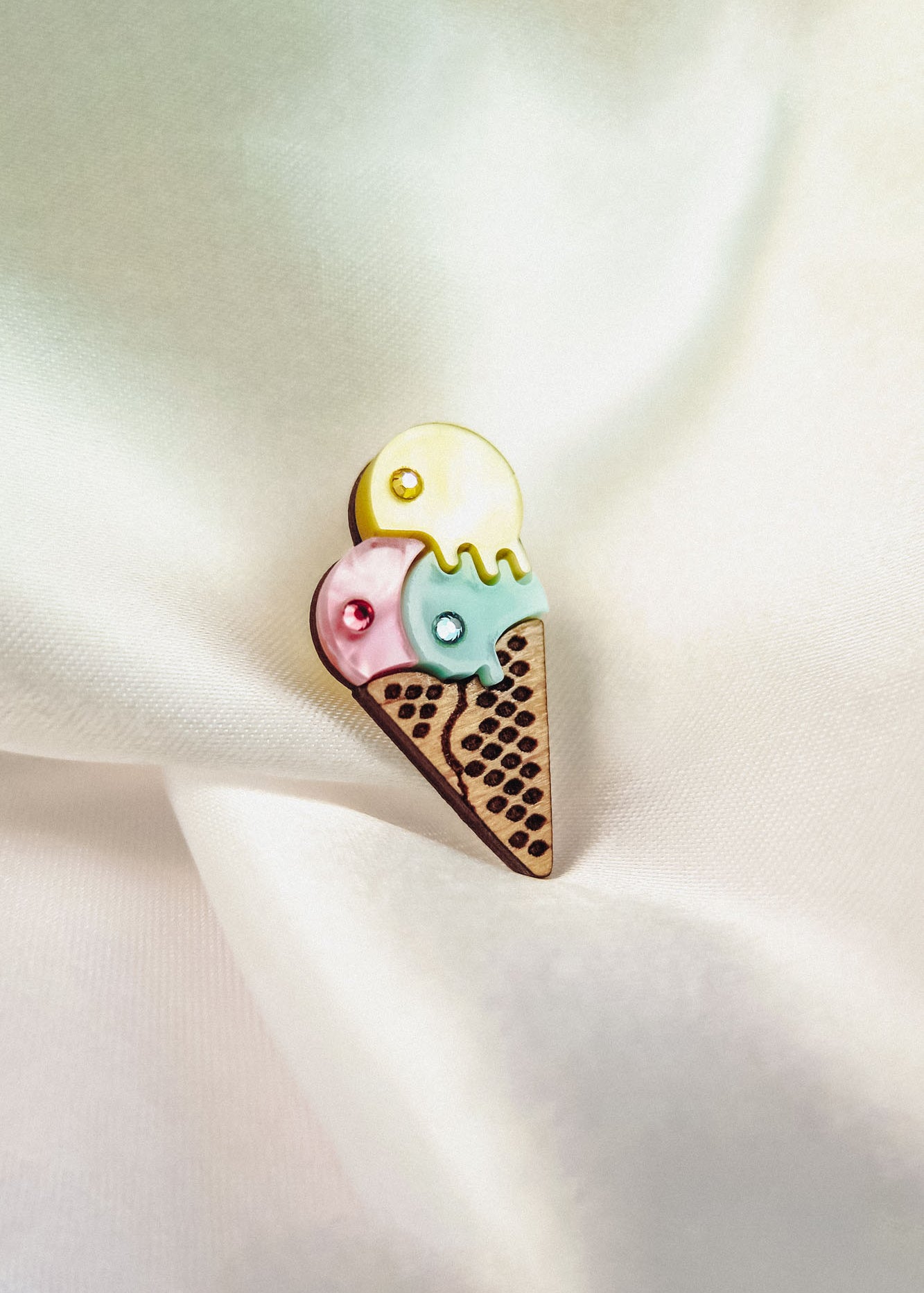 A handcrafted Ice Cream Pin featuring a colorful brooch with three scoops—yellow, pink, and blue—on a waffle cone, resting on soft white satin fabric. Its playful design adds charm to any outfit.