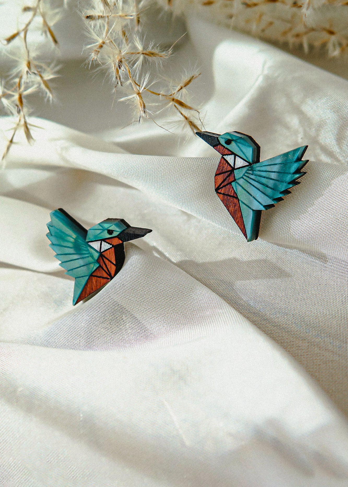 Two colorful, geometric bird brooches, sustainably made and resembling hummingbirds, rest on soft white fabric, with delicate dried plant stems in the background.