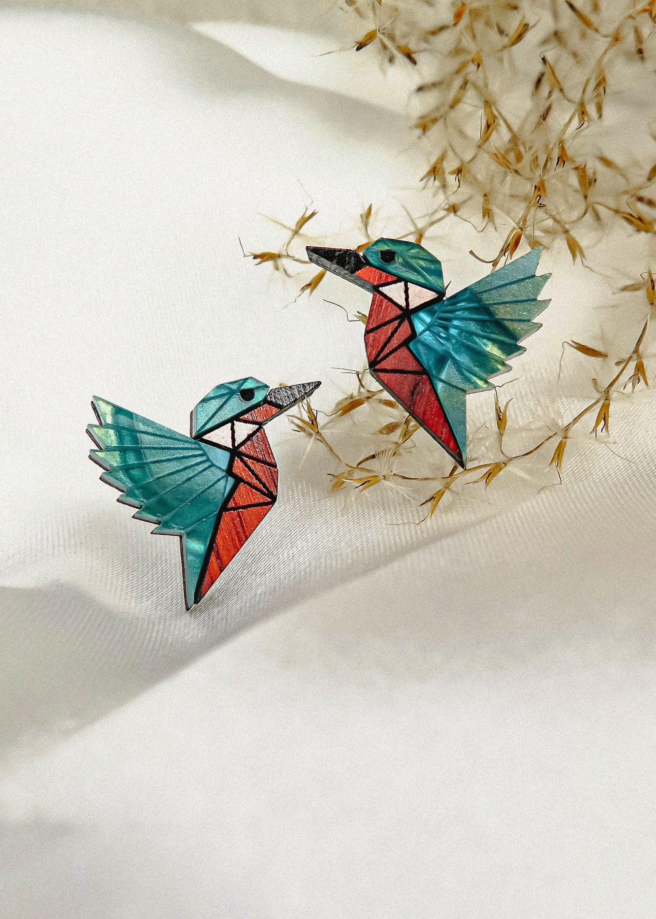 Eco-friendly Kingfisher Stud Earrings: Two colorful origami-style bird earrings with turquoise wings and red bodies, placed on a white textured surface with delicate dried grass in the background.