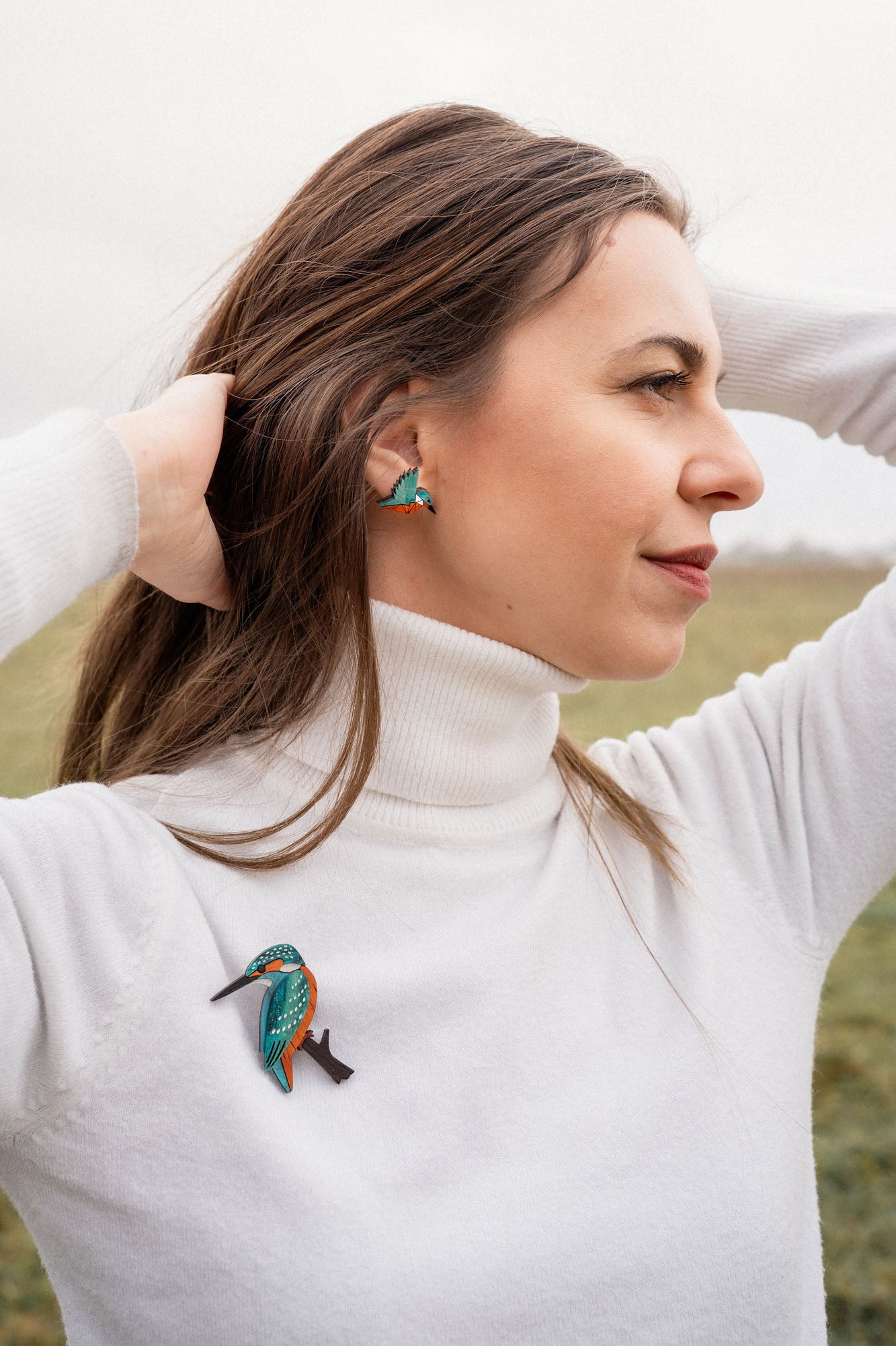 A woman in a white turtleneck stands outdoors with her hands in her hair, wearing matching colorful kingfisher bird earrings and a handcrafted Kingfisher Brooch. The background is a grassy field under a cloudy sky.
