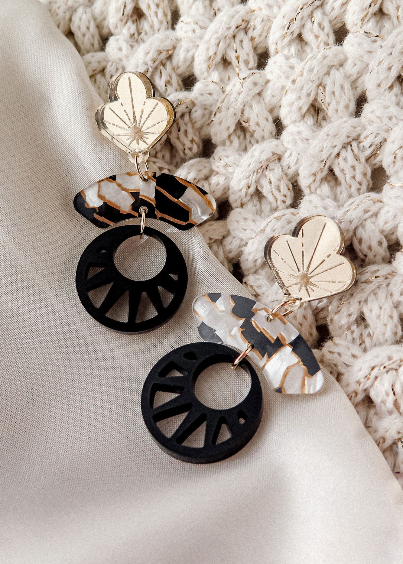 A pair of modern, handcrafted Kintsugi Earrings with gold floral tops, marbled oval shapes, and black circular cutouts, displayed on white fabric next to a textured, cream knitted cloth.