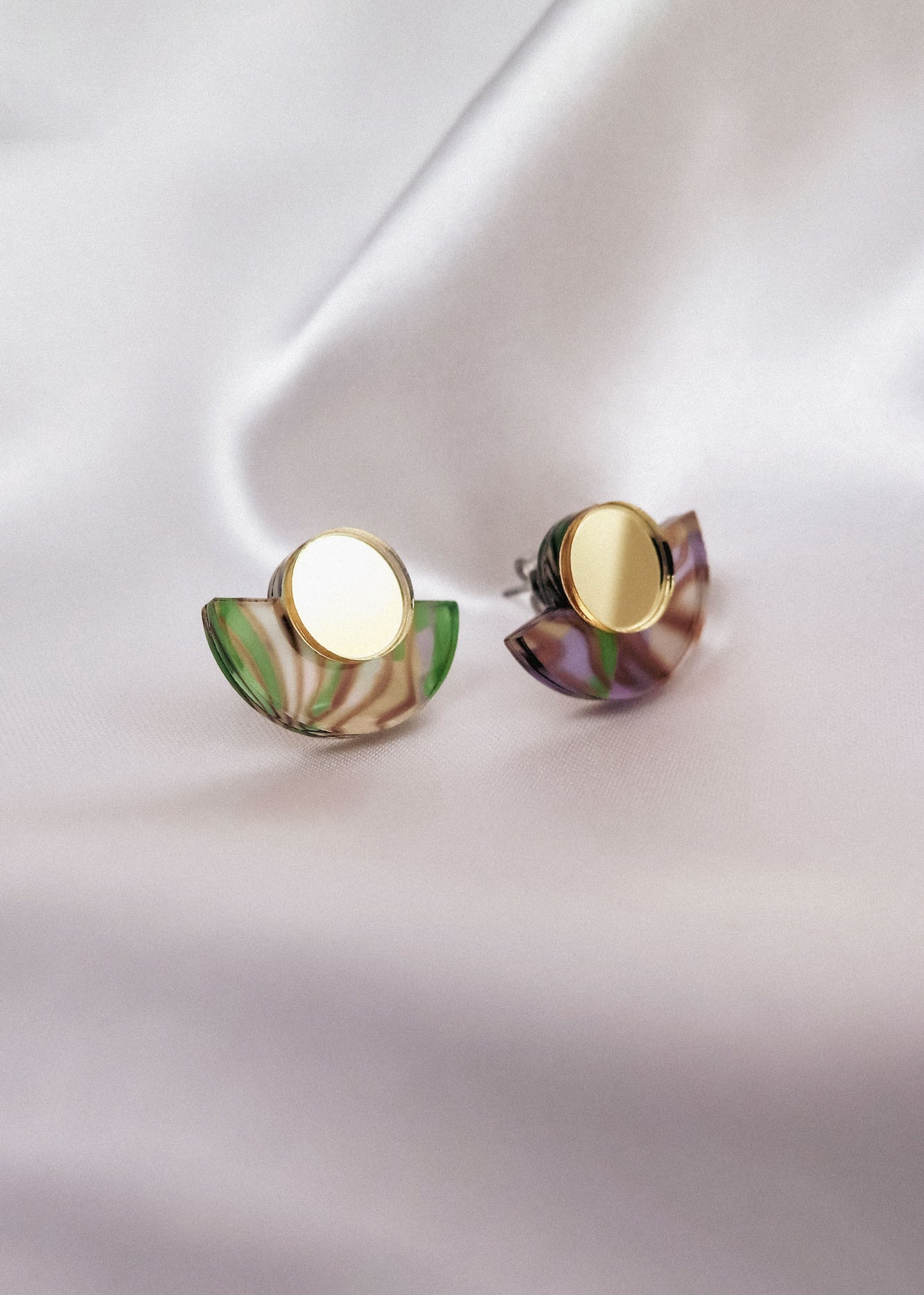 A pair of minimalist geometric stud earrings featuring round gold studs and semicircular marbled green and purple acrylic accents, displayed on soft, white fabric.