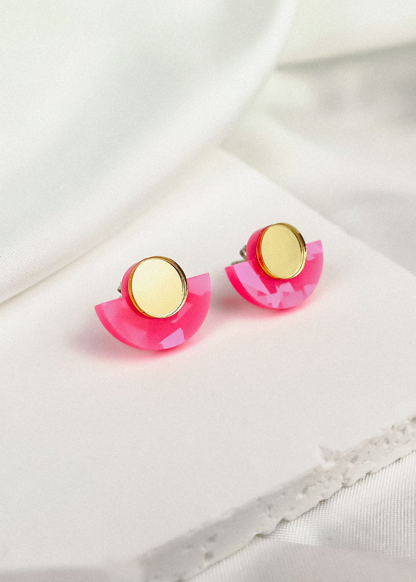 Minimalist Geometric Stud Earrings featuring gold circular tops and pink acrylic semicircles. These lightweight, hypoallergenic studs are displayed on a white fabric surface.
