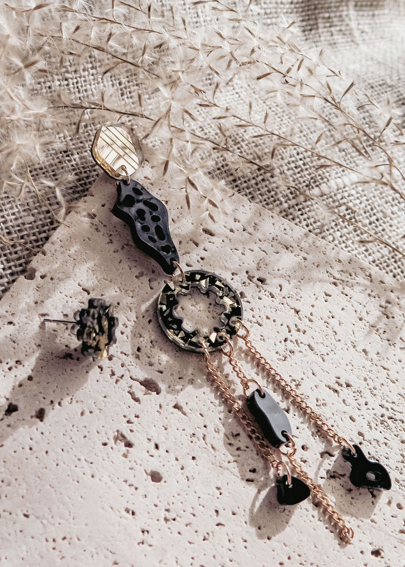 A modern, artistic mismatched extravagant earring in gold, black, and tortoiseshell rests on a textured stone surface, surrounded by dried foliage and beige woven fabric in soft natural light.