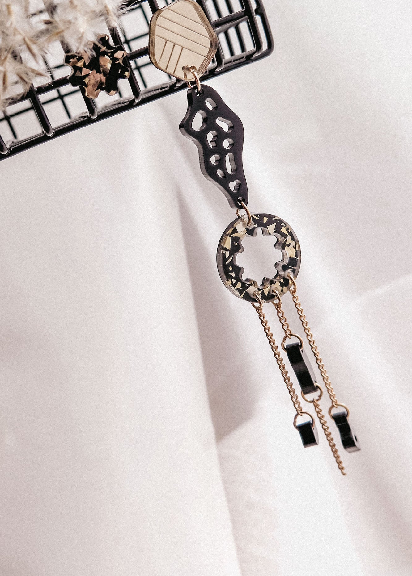 A single handcrafted earring with geometric shapes in gold and black, hanging from a metal rack. This lightweight earring features abstract and circular elements with dangling chains and small black accents.