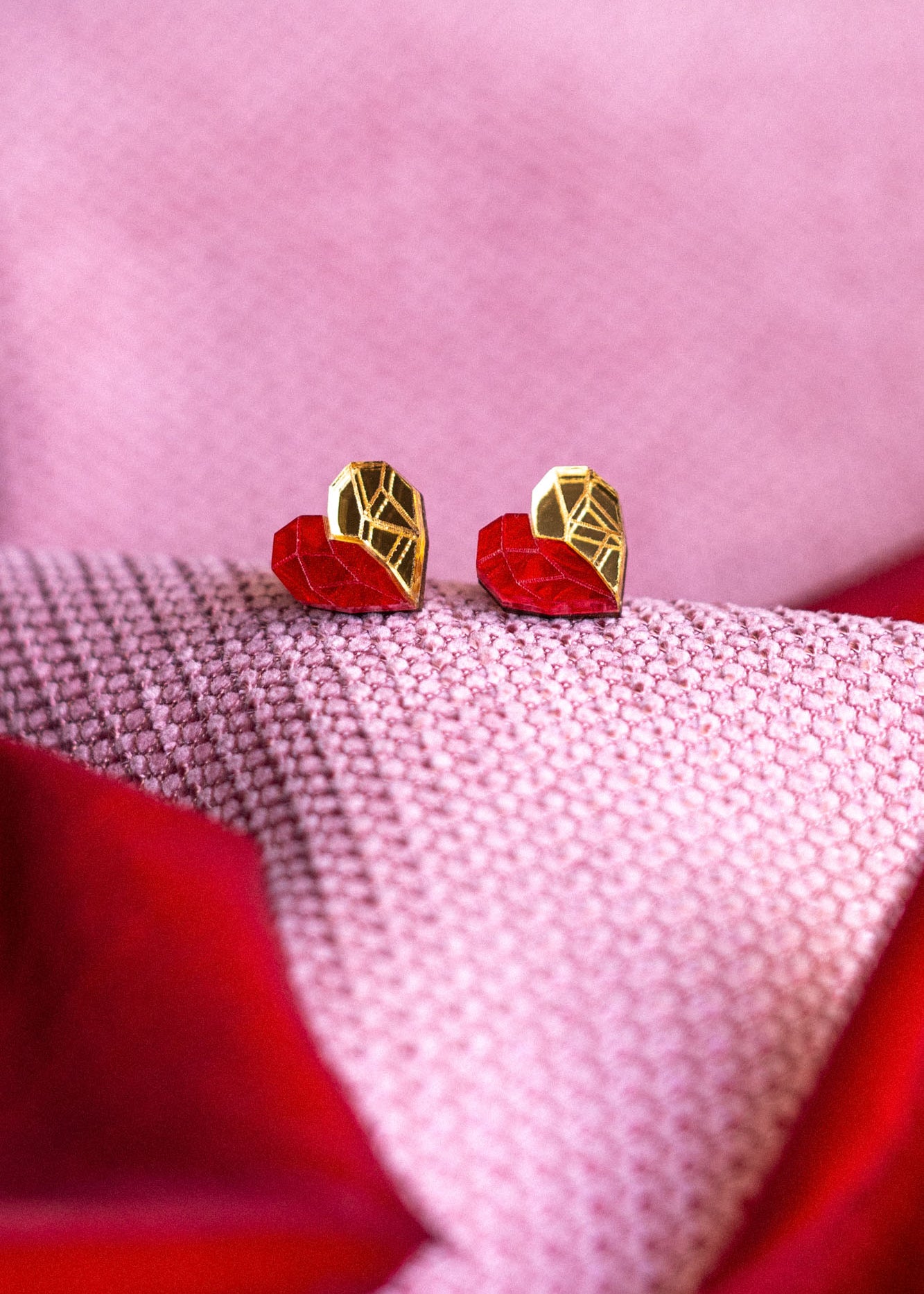 Gold and red geometric heart studs rest on pink and red textured fabric, with the colors and patterns creating a soft, romantic background—a perfect touch of romantic jewelry.