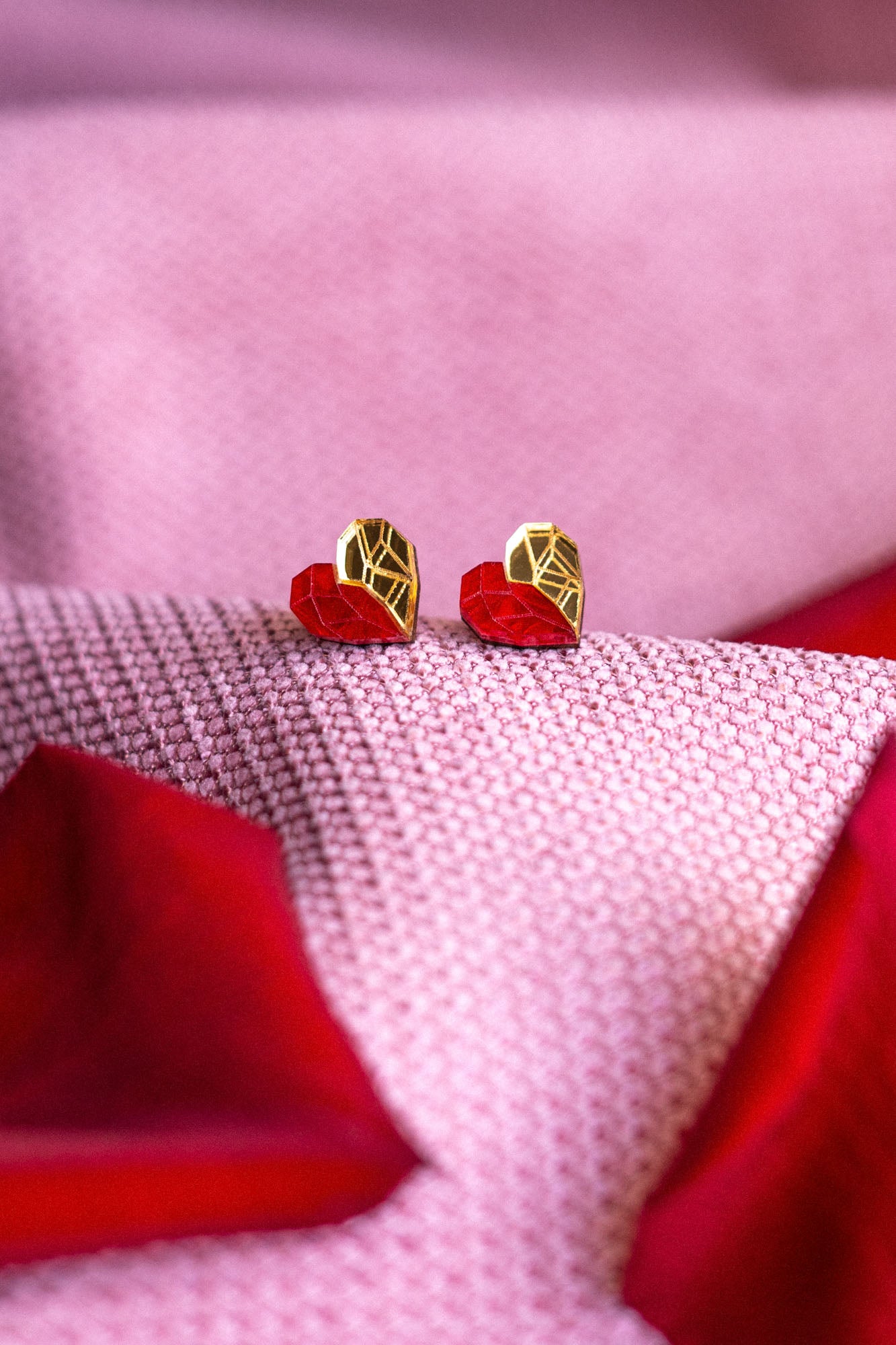 Gold and red geometric heart studs rest on pink and red textured fabric, with the colors and patterns creating a soft, romantic background—a perfect touch of romantic jewelry.