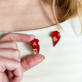 A person with long hair wears a cream top, holding a red and gold geometric One Love Halves Pin shaped like half a broken heart—the other handcrafted half is pinned to their shirt near the collarbone. Perfect as a romantic gift.