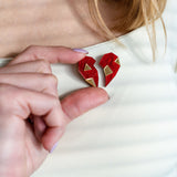 A person wearing a white top holds a red, geometric, handcrafted One Love Halves Pin against their chest. The heart-shaped pin is split into two pieces with gold accents, making it a meaningful romantic gift.