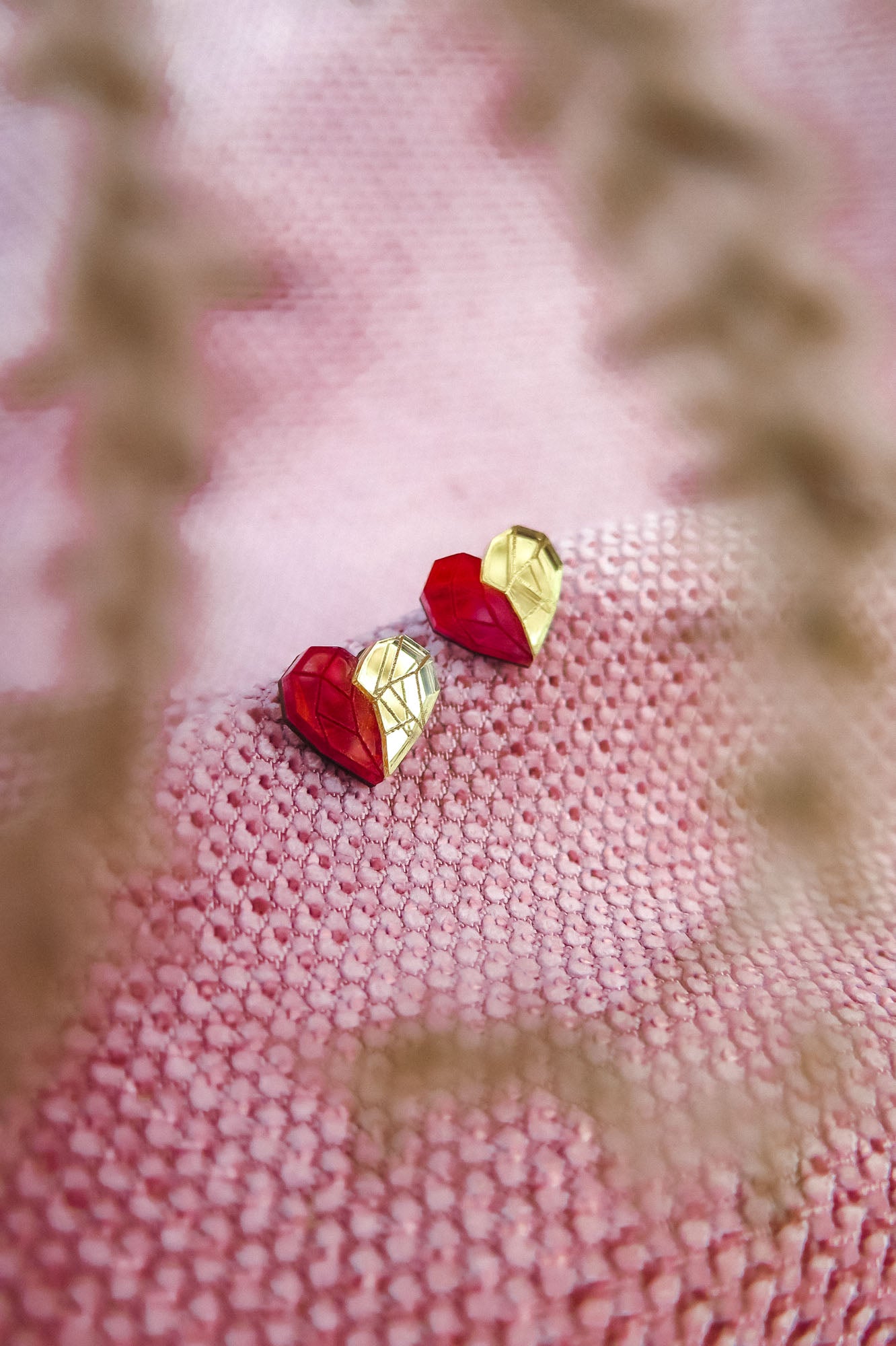 A pair of handcrafted red and gold geometric heart-shaped earrings rest on a textured pink fabric, with blurry plant stems in the foreground—perfect romantic jewelry for any occasion.