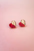 Two origami hearts, each half red and half gold, are placed side by side on a smooth, light pink background, resembling handcrafted earrings or romantic jewelry.
