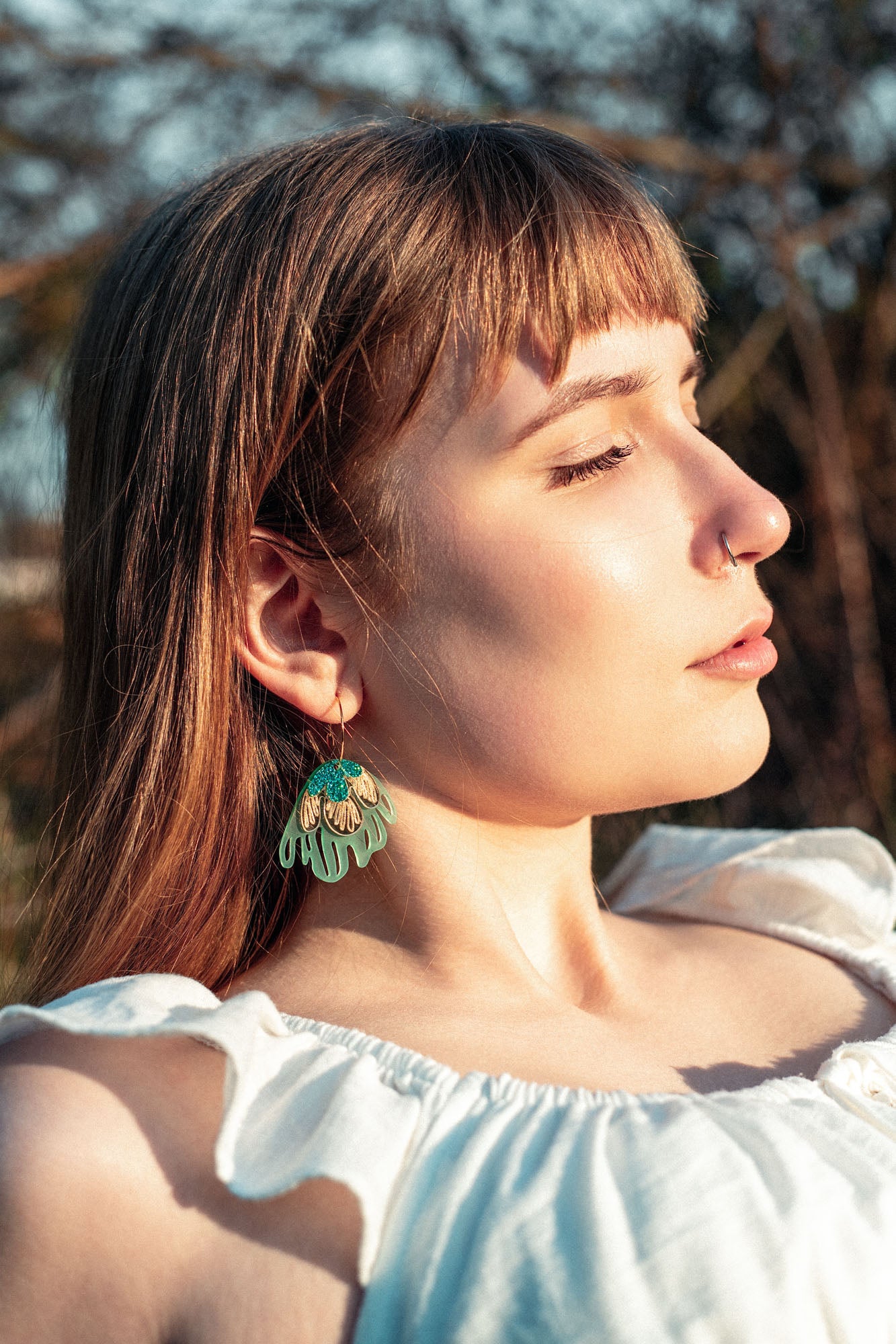 A young woman with light brown hair and bangs, wearing a white top and handcrafted earrings, stands outdoors in sunlight with her eyes closed, appearing peaceful.