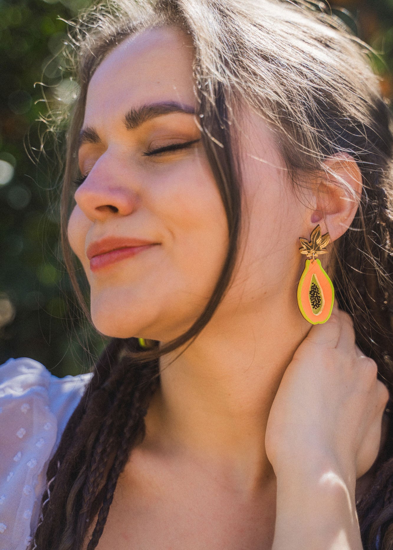 A woman with long brown hair smiles softly with her eyes closed, wearing bright Papaya Earrings made from allergy-free stainless steel and a sheer white top, standing outdoors in sunlight.
