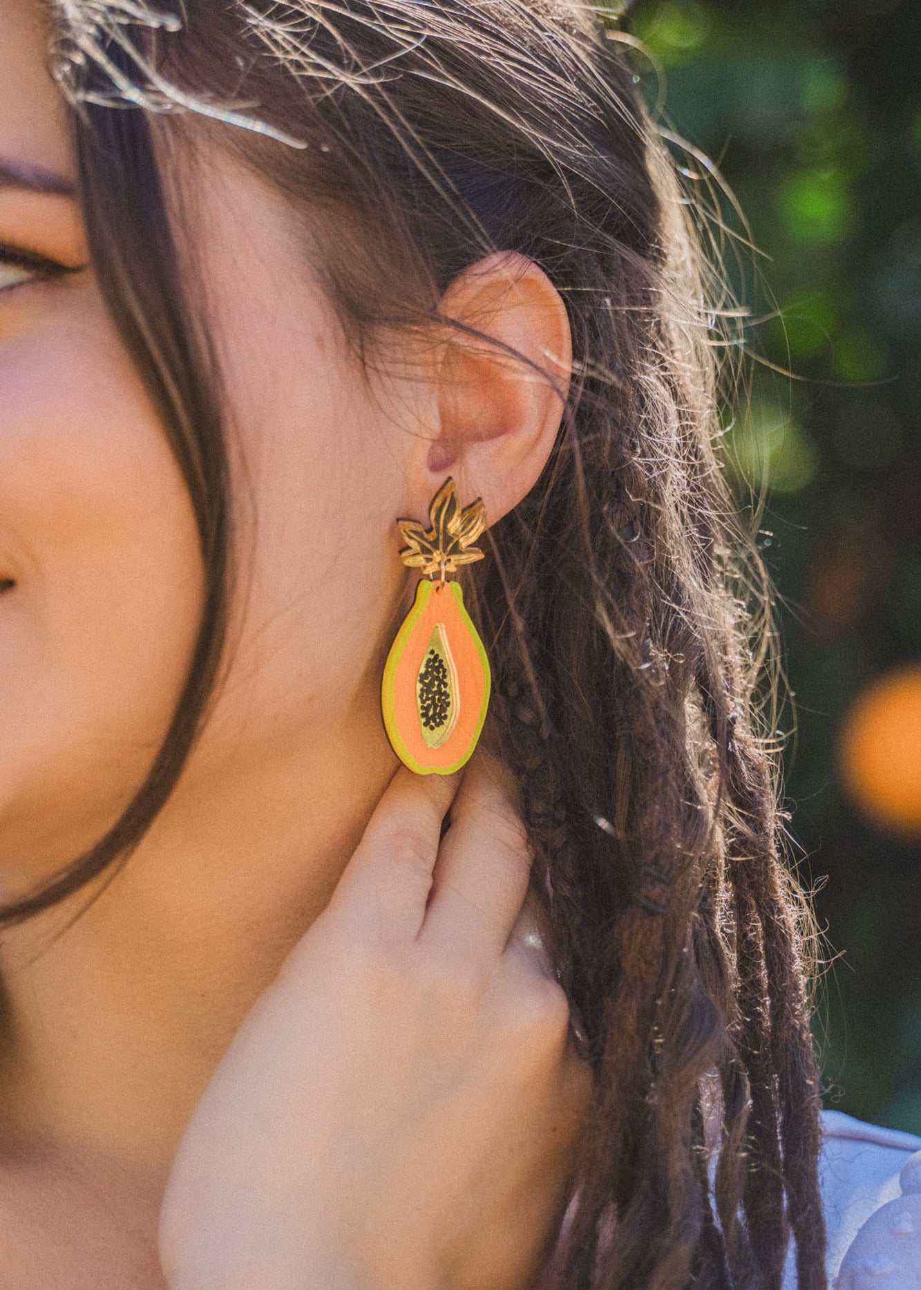 A woman with braided hair wears vibrant Papaya Earrings crafted from allergy-free stainless steel and neon acrylic glass, gently touching one as she stands before blurred greenery.