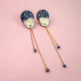 A pair of earrings with blue circular tops reminiscent of the night sky, gold accents, and long gold chains with small blue beads, displayed on a pink background.