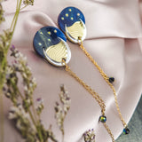 Two round, blue and gold earrings reminiscent of the night sky, with dangling gold chains, are placed on a light pink satin fabric. Small flowers with purple buds frame the foreground, adding to the festival magic of the scene.