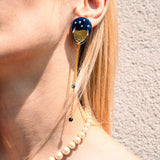 A close-up of a person wearing a blue and gold statement earring with dangling chains that evoke the shimmer of the night sky, paired with a pearl necklace. The person has blonde hair and light skin, and stands against a textured beige wall.