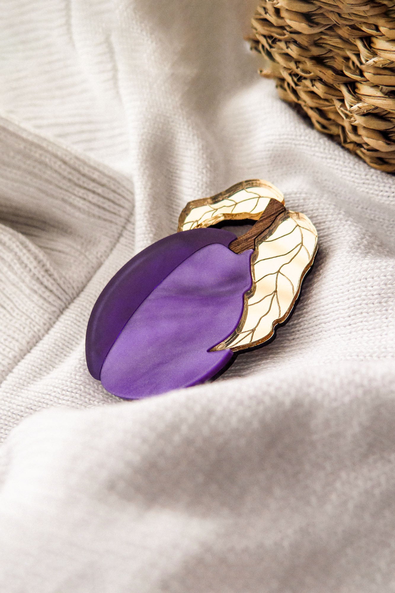 A purple and gold acrylic brooch shaped like a plum with two leaves rests on a soft, white fabric near a woven basket.