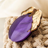A purple brooch shaped like a plum with gold-colored leaves, handmade from recycled wood, rests on a knitted beige fabric.