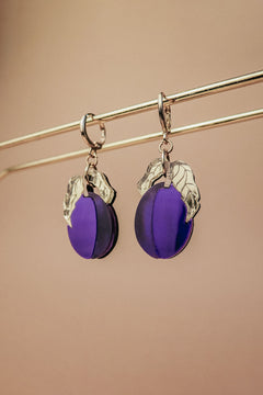 A pair of elegant purple plum earrings with silver leaf accents hang from a gold-colored rod against a beige background.
