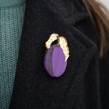 A person wearing a dark coat with an elegant brooch shaped like a purple fruit and two gold-colored leaves pinned to the lapel. The person is also wearing a teal knit sweater.