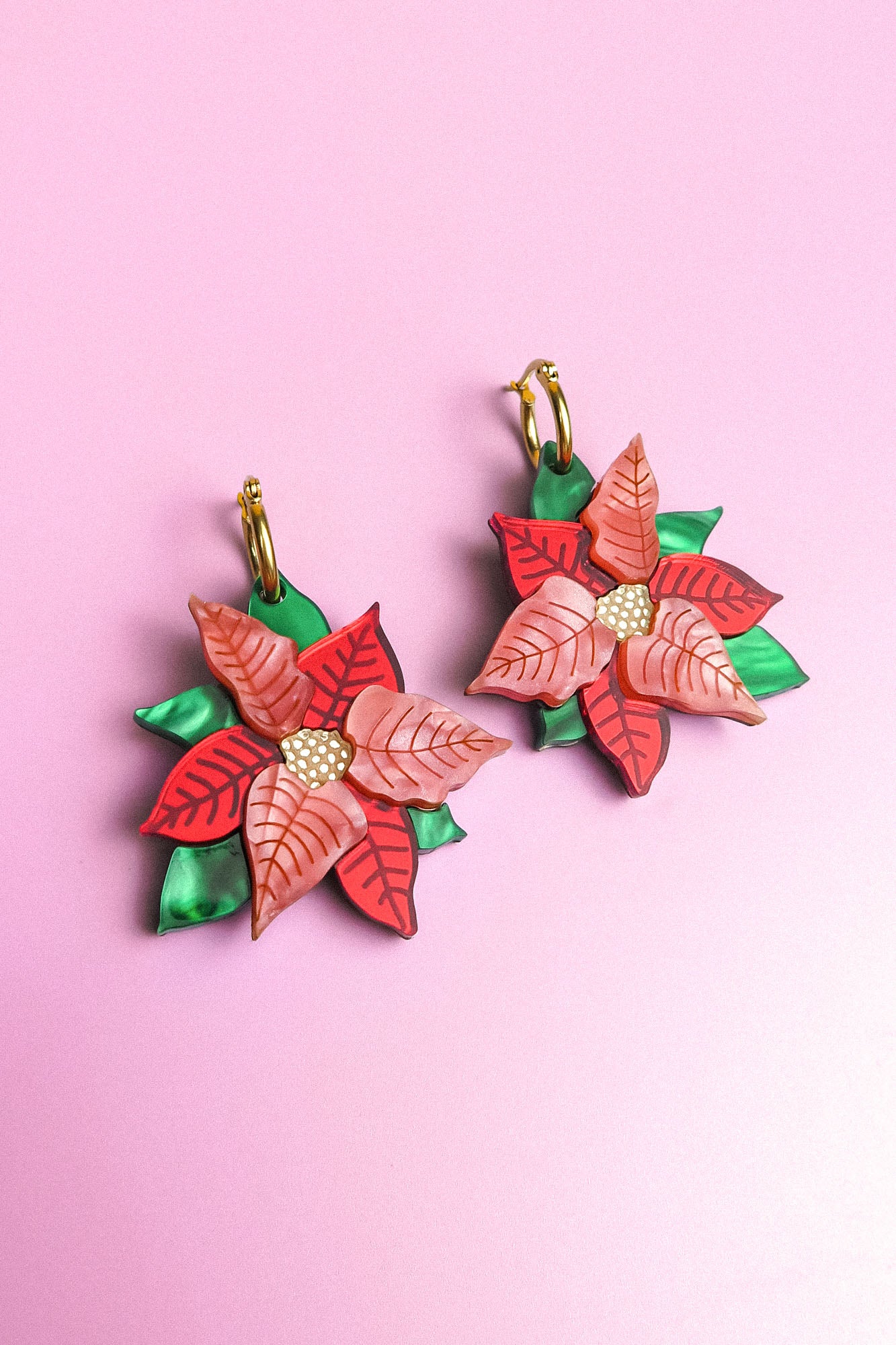 A pair of Poinsettia Statement Hoops shaped like poinsettia flowers with red and pink petals, yellow centers, and green leaves, displayed on a light purple background—perfect festive earrings for your holiday jewelry collection.